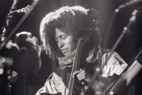 In a spotlight, a Black woman plays the cello amongst an orchestra.