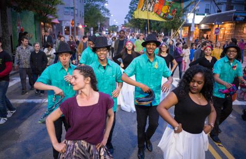 People dance on the street in Boston's Latin Quarter. Men in matching teal tees and black hats dance behind the first row of people.