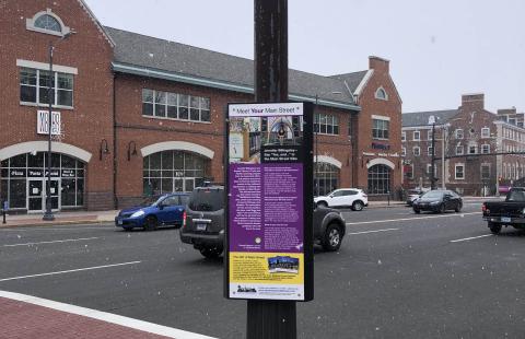 Image of a sign on a lamp post on a sidewalk with the town center in the background.