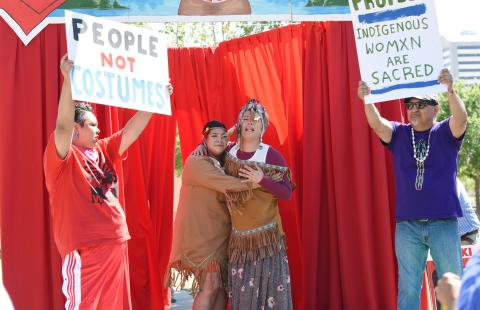Two women, in native garb, embrace while two men hold up signs that read "people not costumes" and "indigenous womxn are sacred."