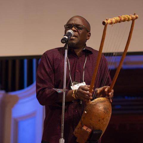 artist Samite Mulondo plays a litungu, a seven stringed instrument, and sings into a standing mic at the 2018 Idea Swap on the Great Hall stage at Mechanics Hall. Samite's eyes are closed as he performs. Blue lighting on the stage and a blank projector screen are seen in the background