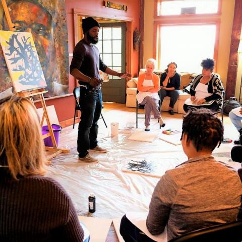A Black man holding a paint brush with a canvass behind him and surrounded by a semi-circle of seated students.