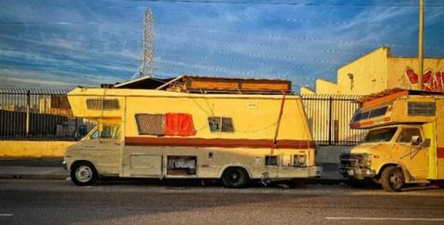 An RV in disrepair parked on the side of a road. A tall fence and radio tower appear in background, with blue sky and late day sunlight reflecting on the vehicle.
