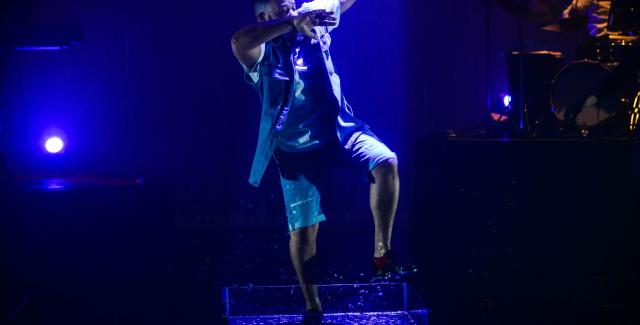 On a stage, a Black man lifts his leg and raises both arms to the right and across his face.