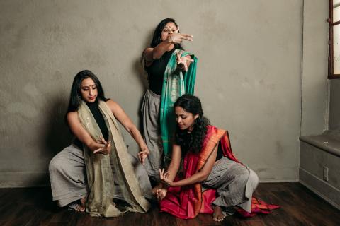 Three Indian women form a triangle and move their hands in a room with tan walls.