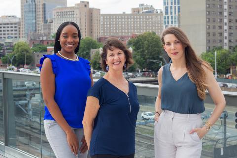 Indira is a Black woman with long black hair and she wears a blue blouse. Cheri is a white woman with short brown hair and she wears a navy, v-neck. Kristin is a white woman with long brown hair that has blonde highlights and she wears a stone v-neck with khakis. They all pose in front of the Boston skyline.