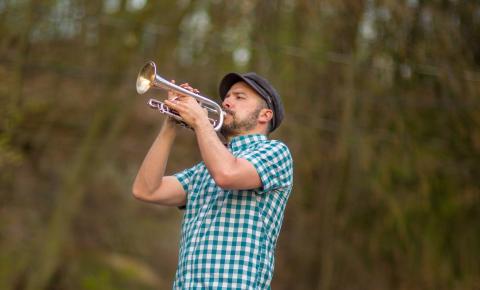 A man in a plaid shirt playing the trumpet outside with trees in the background