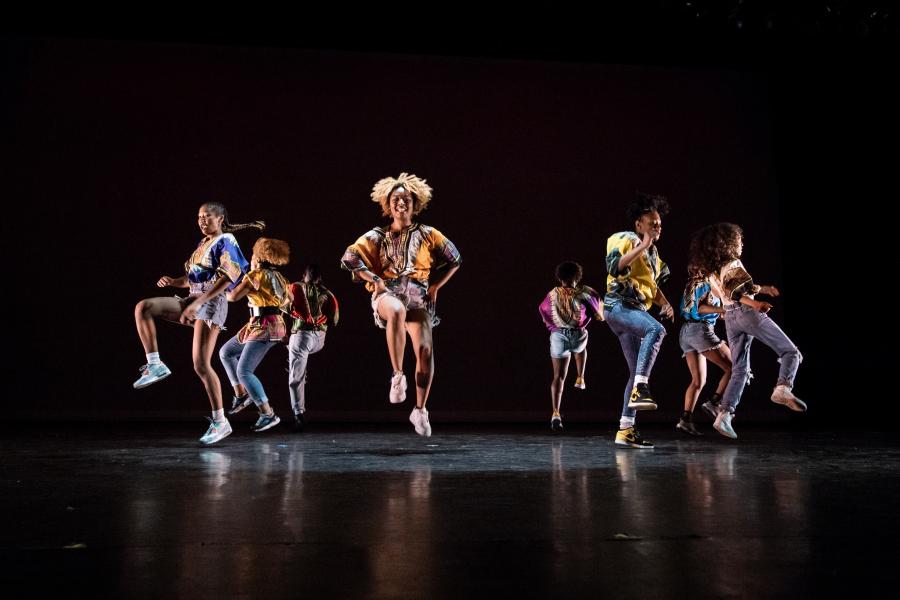 On a stage with black curtains, eight Black dancers, in streetwear, lift their right leg with their knees bent.