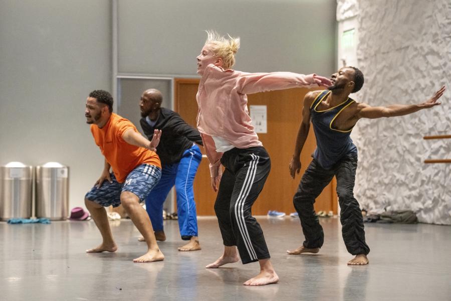 In a studio space, four dancers, three Black men and a white woman, throw their arms back.