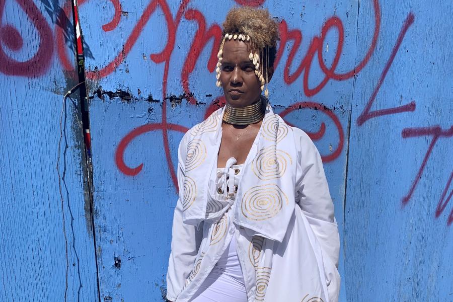 A Black woman, in all white with a gold swirl pattern, poses in front of a blue wall with graffiti.