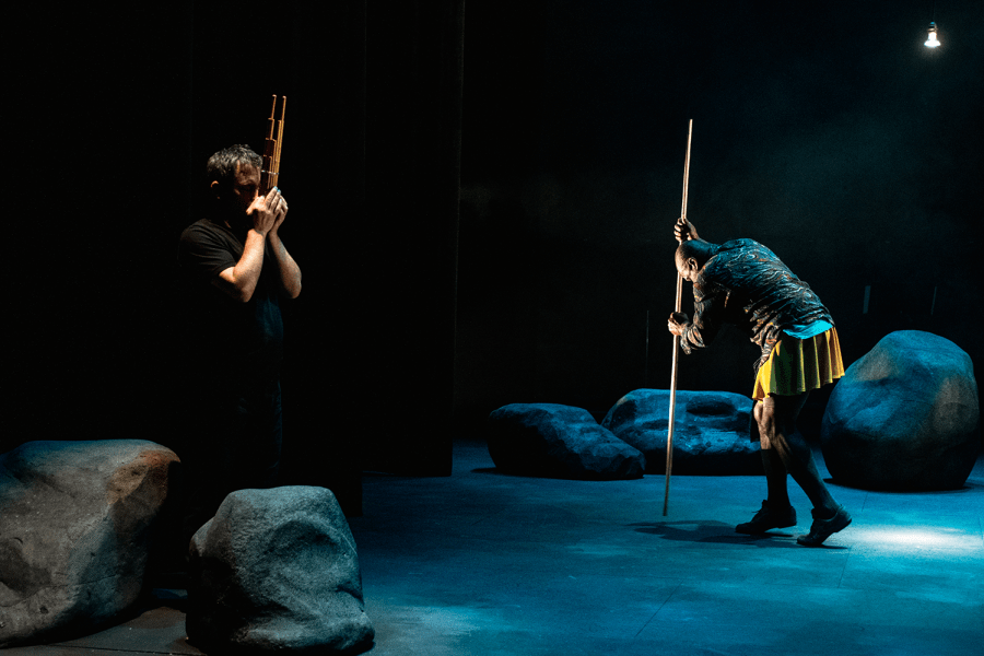 On a stage, a man holds a stick next to his face and a woman leans over another stick by some rocks.