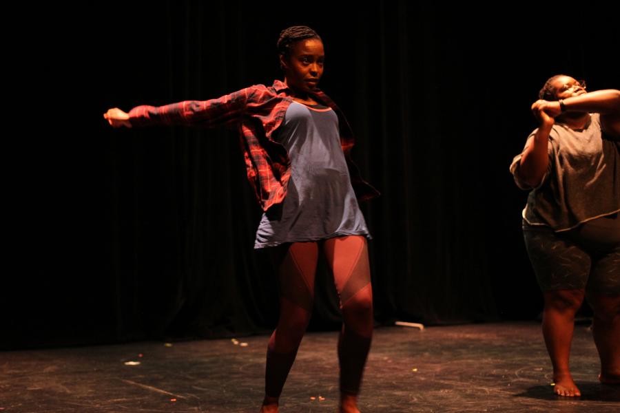 Two Black women dance. One wears cream sweats and another has a denim romper and a red plaid button down.