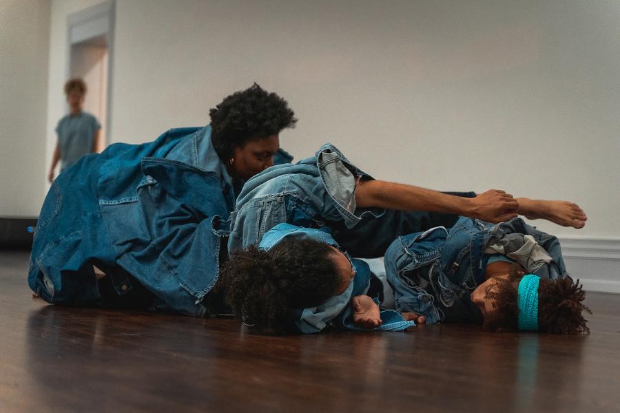 Inside, three black women, wearing denim, roll around on each other on the floor.