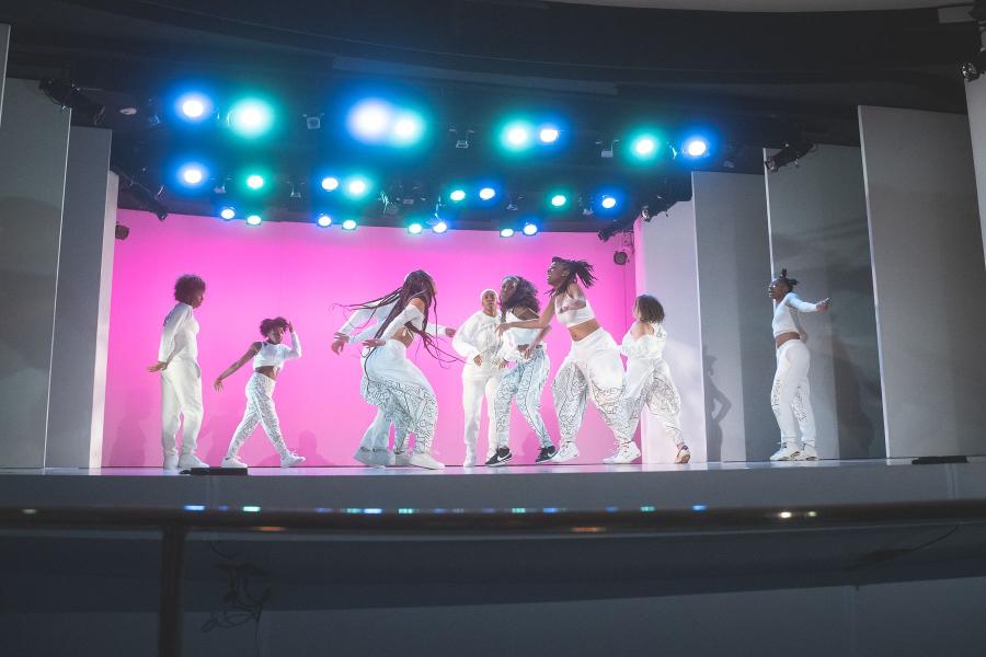 Black women, in white outfits, are divided in half and face each other, in front fo a pink backdrop.