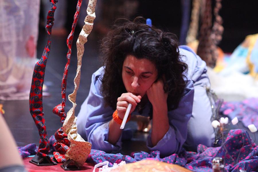 Next to flowers made of fabric, a woman lays on the ground blowing into a straw.