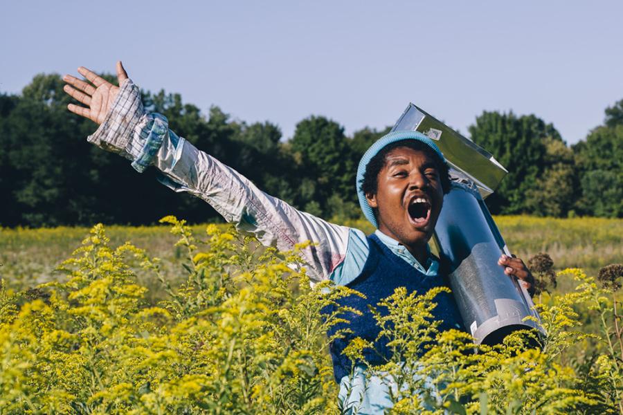 A young Black person carries something shiny in a field of flowers and screams.