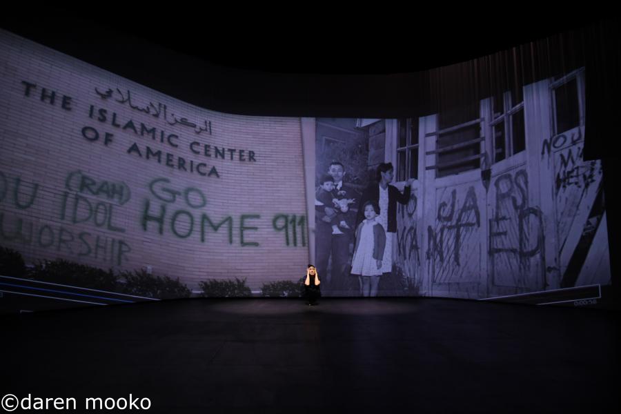 A woman crouches in front of a projection of old photos. On the old photos are the words or phrases ("Go home" and "911") spray painted on the side of the Islamic Center of America.
