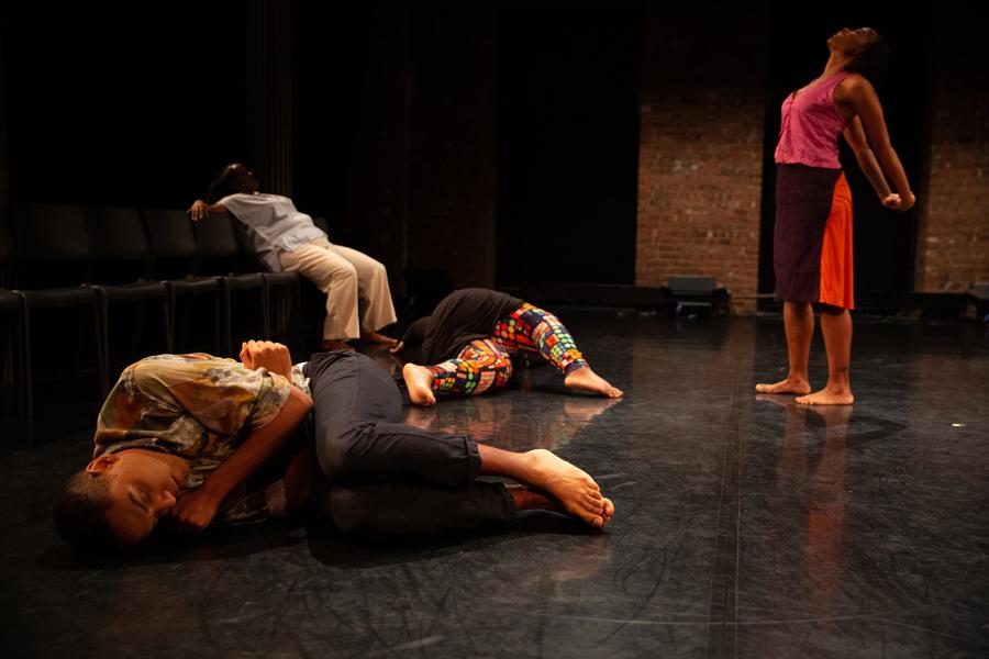 On a stage, four folks perform; three are lying down and one is standing an stretching her arms.