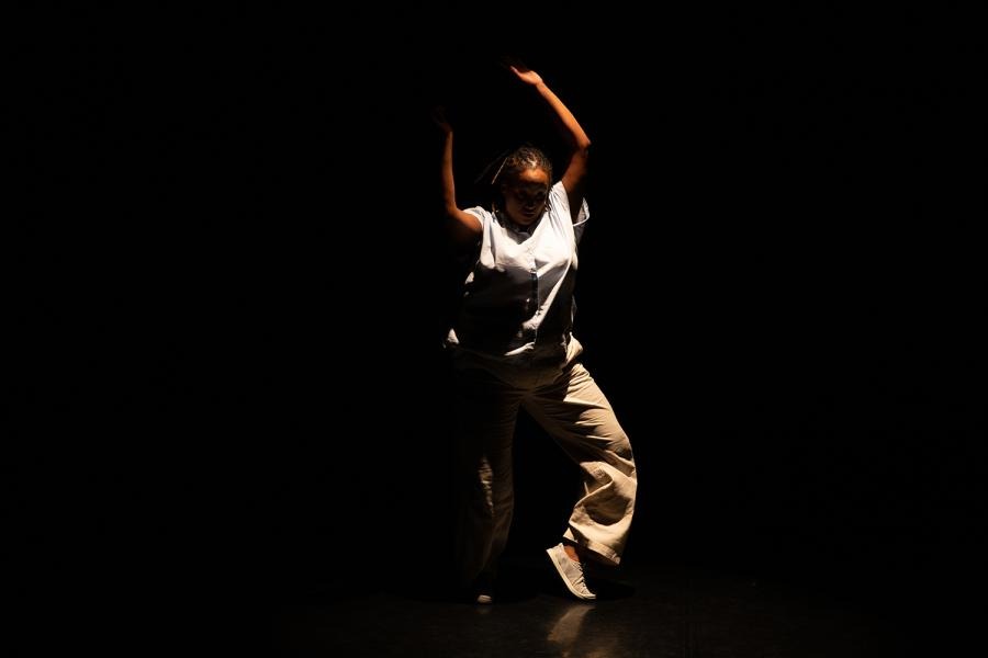 In a spotlight, a Black woman, in khaki, spins with her arms up.