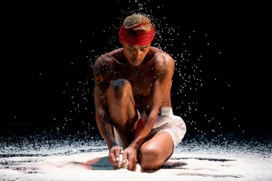 A brown man, with blonde hair in a red wrap, kneels in salt that falls from the ceiling.