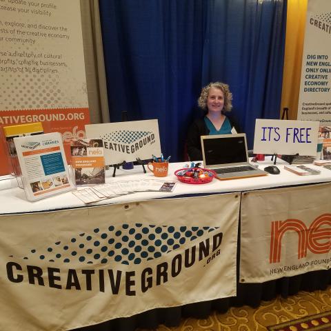 A woman stands behind two tables. One has a CreativeGround logo banner and another has a NEFA logo banner.