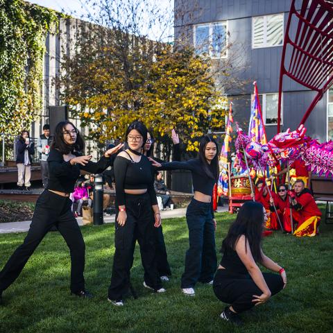A group of four young, Asian girls dance in all black. There's a sculpture made of red metal behind them and folks performing a dragon parade.