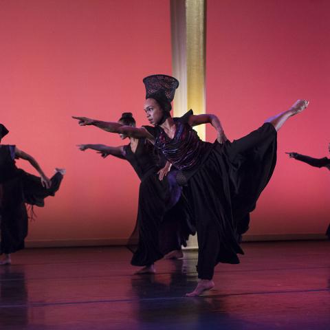 A dancer, in an ensemble of dancers, lifts her leg back and up and points forward.