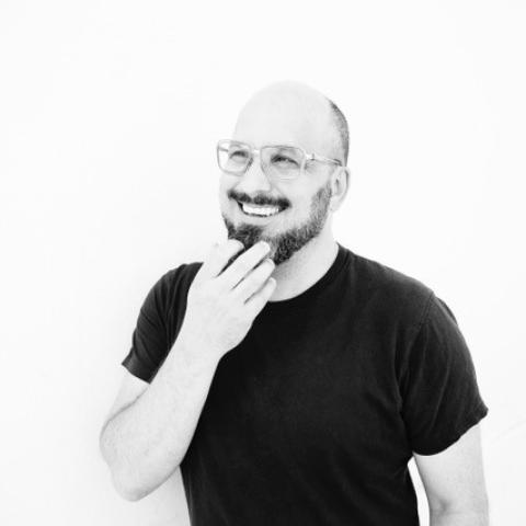 A black and white photo of a white man of mixed Euro-heritage, beard, bald head, and glasses. He is wearing a black shirt, and he is touching his chin with a big smile.