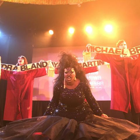 A woman in black sparkly evening wear on stage in front of people in red robes holding banners with names written on them