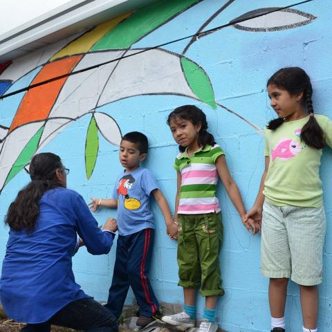 A woman leans down to help a young boy who leans against a mural with two young girls.
