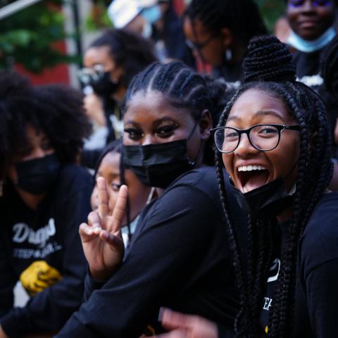 Outside, three young Black girls pose and smile together, while wearing black clothing, face masks, and eyeglasses.