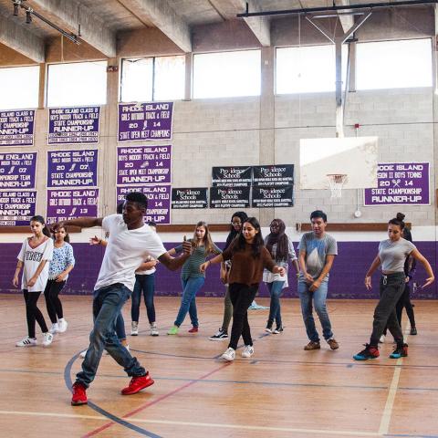 In a high school gymnasium, a man in a white tee leads a group of students in a dance.