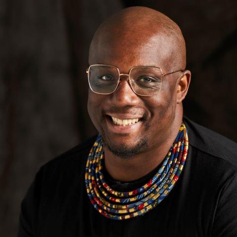 A Black man in thin framed glasses smiles. They have a goatee and wear a layered necklace.