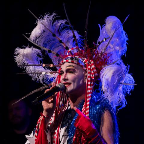 The artist holding a microphone and wearing an elaborate red, white, and blue costume with feathers, stars, and sparkles.