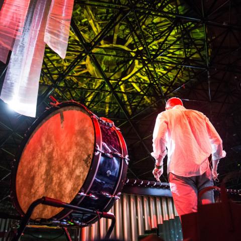From behind and below, a man stands next to a large drum, amid colorful spotlights.