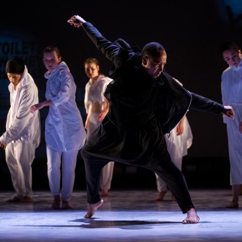 A man in black dances in the foreground while dancers in white dance in the background.