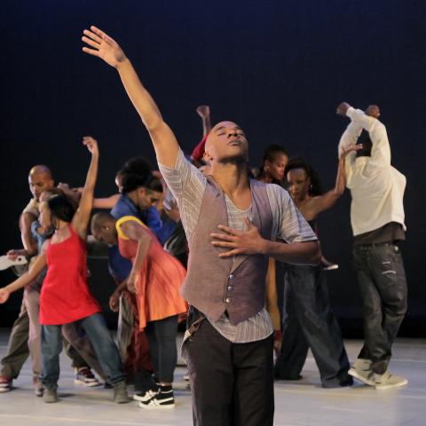 A man in a vest throws his arm up in the foreground and in the background a group of dancers move in a circle.