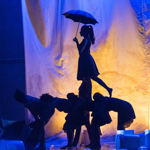 A woman with an umbrella walks over the backs of other dancers leaned over. They're back lit by a yellow lamp in a blue space.