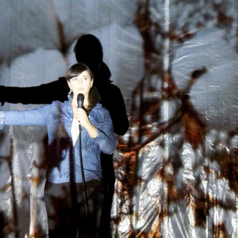 Lit by a projection of wheat, a woman holds her arm out while wearing a jean jacket.
