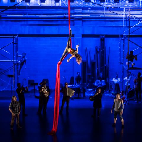A person dangles from a red fabric on a stage; musicians stand around it. 