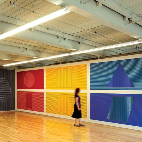 An indoor gallery space, folks look at large scale murals in color blocked designs.