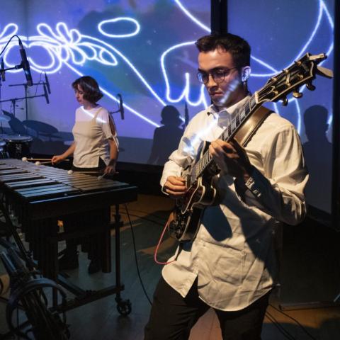 A white man plays the guitar and a white woman plays the piano. A projection of white lines over purple and blue swirls behind them.
