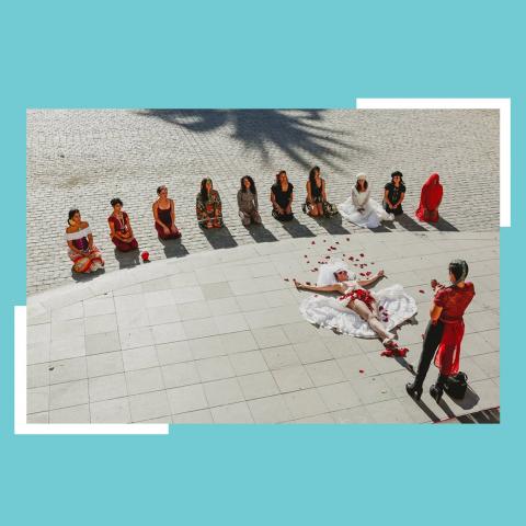 Twelve women, kneeling in a line, watch as a woman lies on the outdoor stone ground and another woman throws petals over her body. They're all in reds and whites.