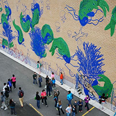 A crowd looks at a wall covered in blue and green graffiti made of tape.