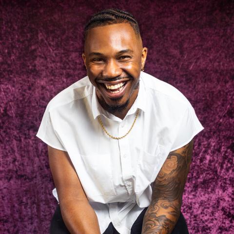 Tariq is a Black man in a white sleeveless shirt. He has a sleeve of tattoos and torn black jeans. He poses in front of a purple velvet fabric.