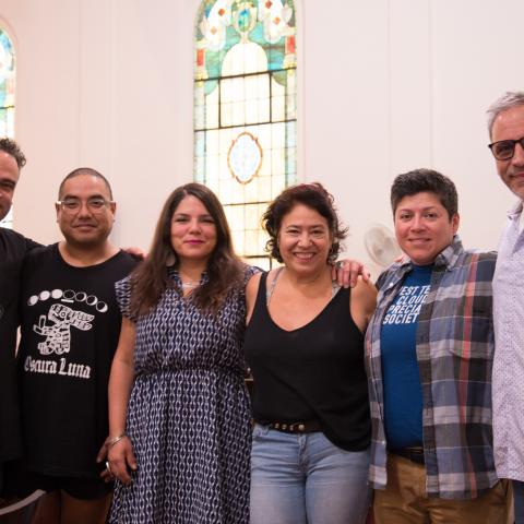 Six people in casual clothing standing arm in arm facing the camera. Large stained glass windows are in the background.