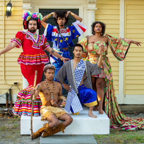 Five folks, in ornate and colorful costumes, pose in front of a yellow house.