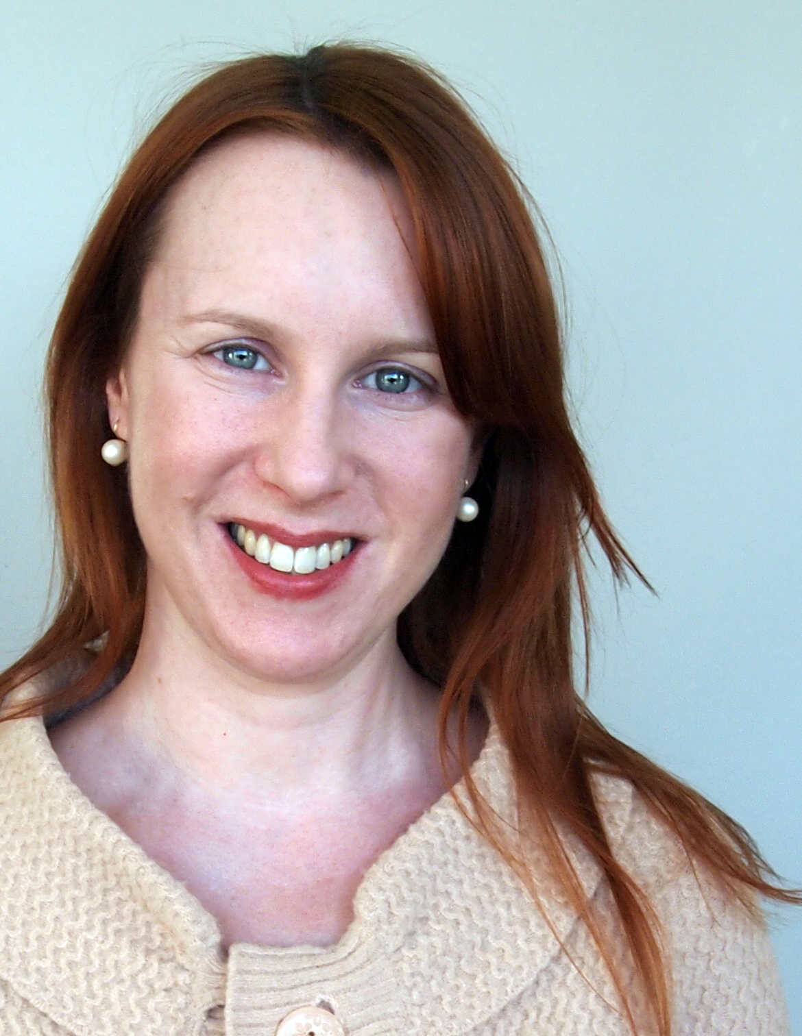 Sara smiles in front of a plain backdorp in a tan sweater