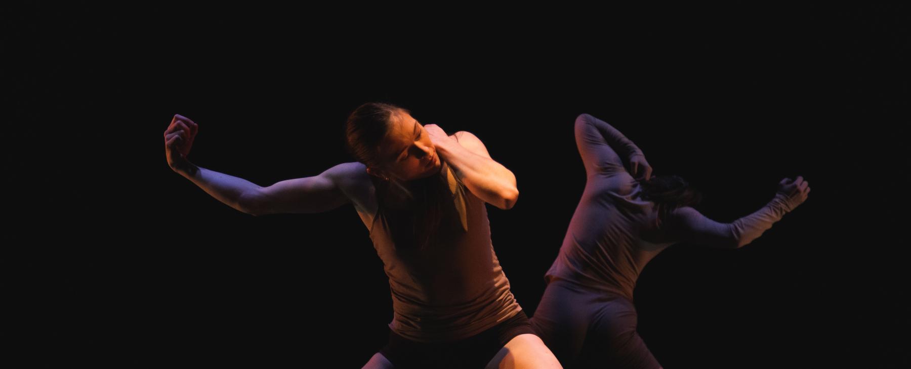 Two dancers mirror each other's movements, with the dancer in the foreground in a spotlight.