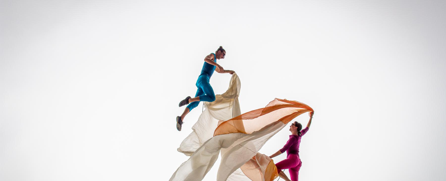In a white void, two dancers (in brightly colored costumes) dance with orange and beige fabric.